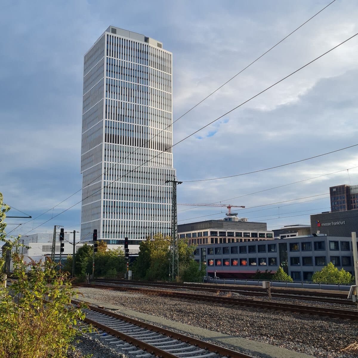 Ein moderner, hoher Wolkenkratzer mit heller Fassade ragt hinter Bahngleisen und Oberleitungen in den Himmel, im Hintergrund sind weitere Gebäude zu sehen.
