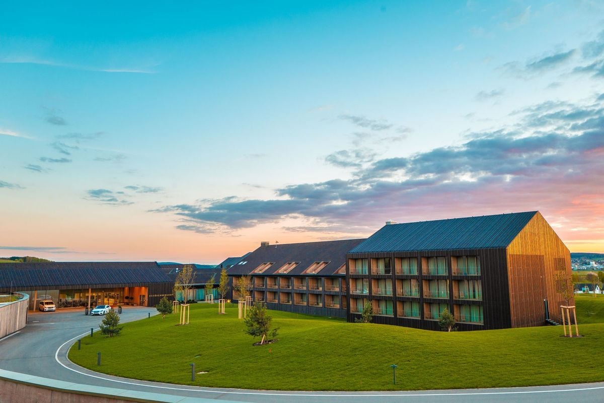 Ein modernes Hotelgebäude mit dunkler Holzfassade und Satteldach steht in einer hügeligen Landschaft vor einem farbenfrohen Abendhimmel bei Sonnenuntergang.