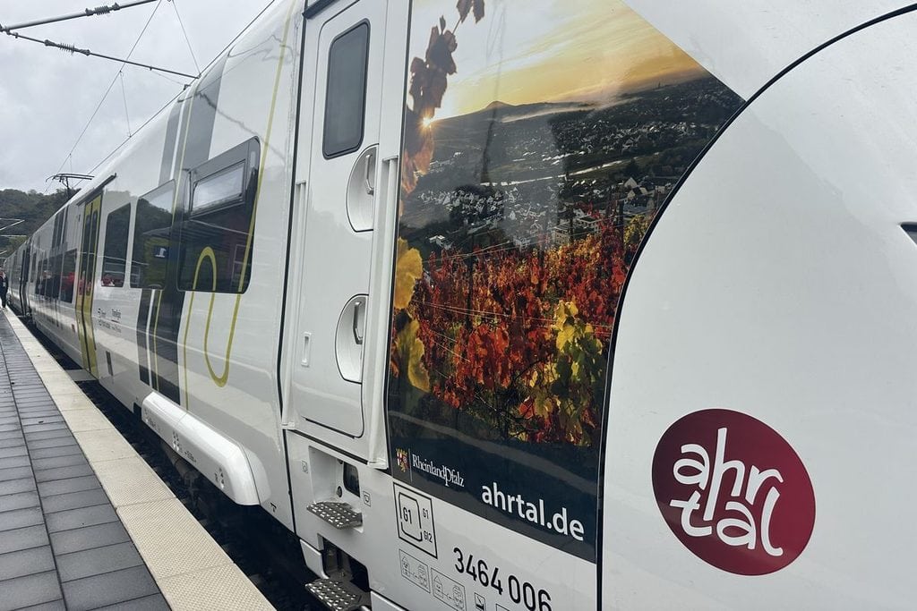 Ein weißer Regionalzug steht an einem Bahnsteig, dessen Außenwand mit einem großflächigen Foto von herbstlichen Weinbergen und dem Ahrtal bedruckt ist.