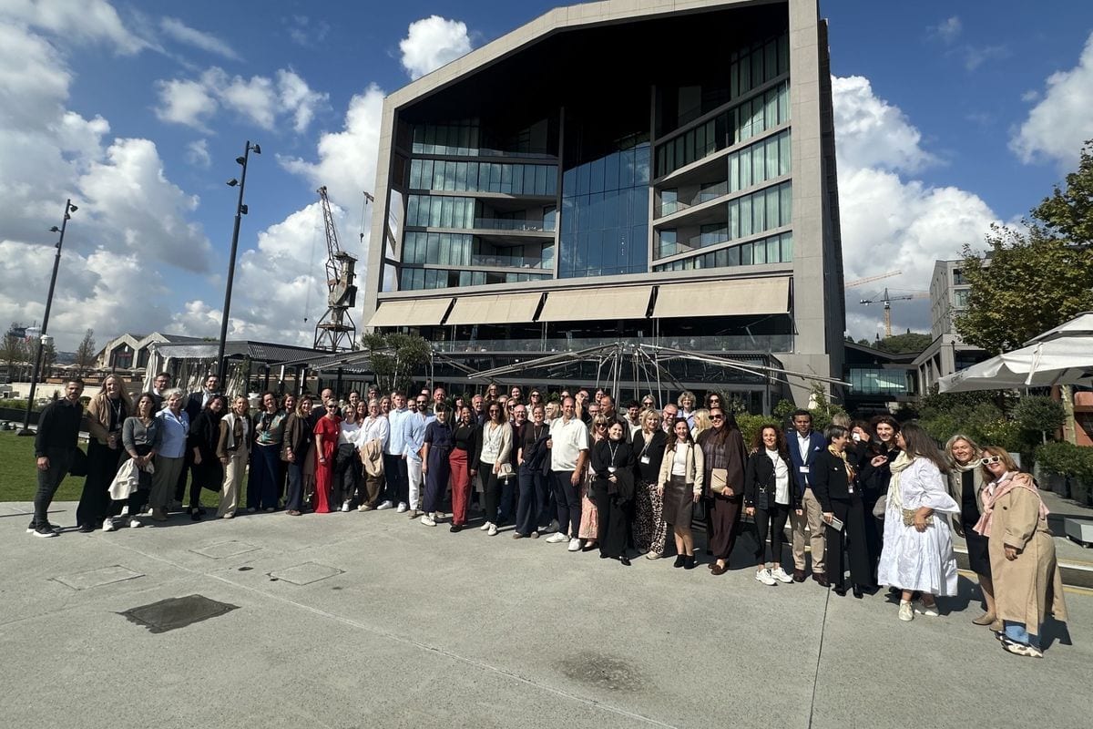Eine große Gruppe von Menschen steht auf einem gepflasterten Platz vor einem modernen, hohen Hotelgebäude mit Glasfassade unter einem blauen Sommerhimmel.