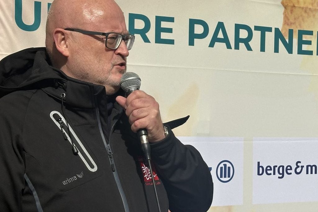Ein Mann mit Glatze und Brille spricht in ein Shure-Mikrofon. Er trägt eine schwarze Funktionsjacke vor einem hellen Hintergrund mit Firmenlogos im B2B-Stil.