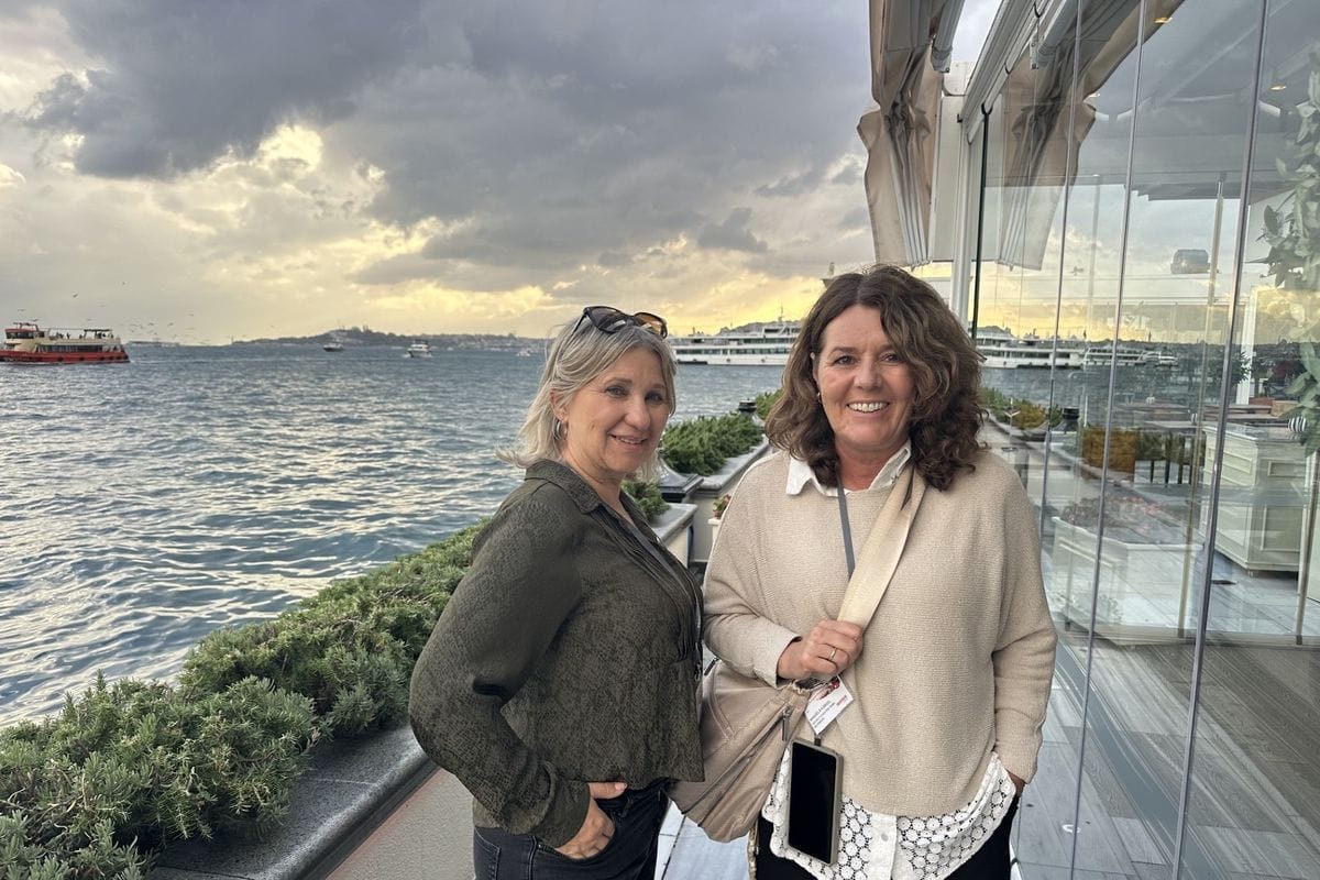 Zwei Frauen stehen lächelnd auf einer Terrasse am Wasser mit Blick auf den Bosporus, im Hintergrund sind Schiffe und ein bewölkter Himmel zu sehen.