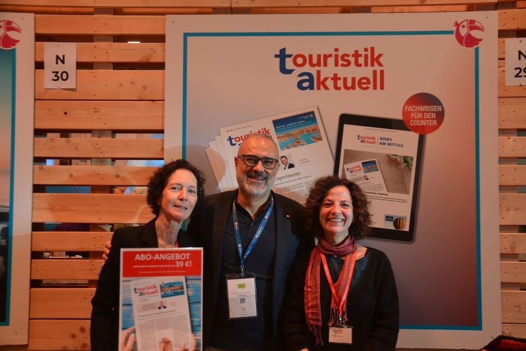 Drei Personen stehen lächelnd vor einer Werbewand von touristik aktuell. Eine Person hält ein Schild mit einem Abo-Angebot in die Kamera. Messeumgebung.