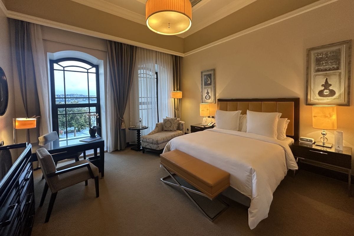 Ein elegantes Hotelzimmer mit einem großen Doppelbett, einer Sitzbank am Fußende, einem Schreibtisch und einem Fenster mit Blick auf eine Wasserfläche.
