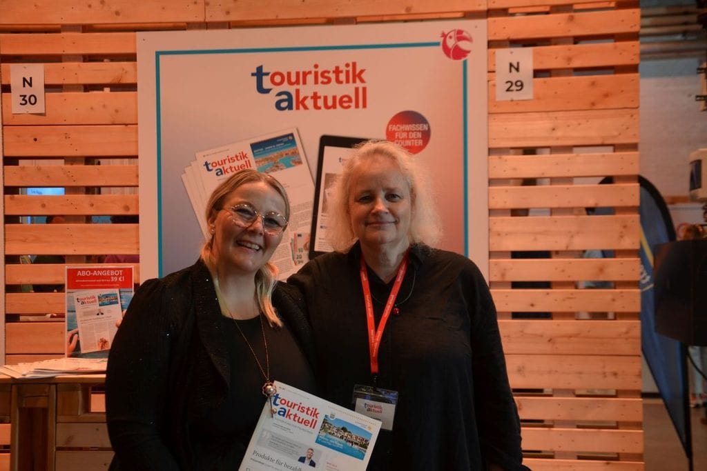 Zwei Frauen stehen lächelnd vor einem Messestand mit dem Logo von touristik aktuell, im Hintergrund ist eine Wand aus hellen Holzlatten zu sehen.