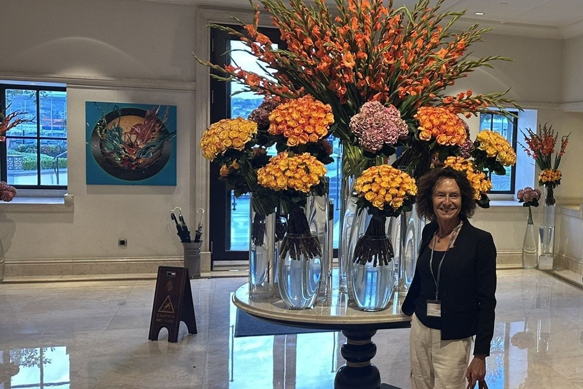 Eine Frau mit dunklem Haar steht in einer hellen Hotellobby neben einem großen, runden Tisch mit üppigen Blumenarrangements in hohen Glasvasen.
