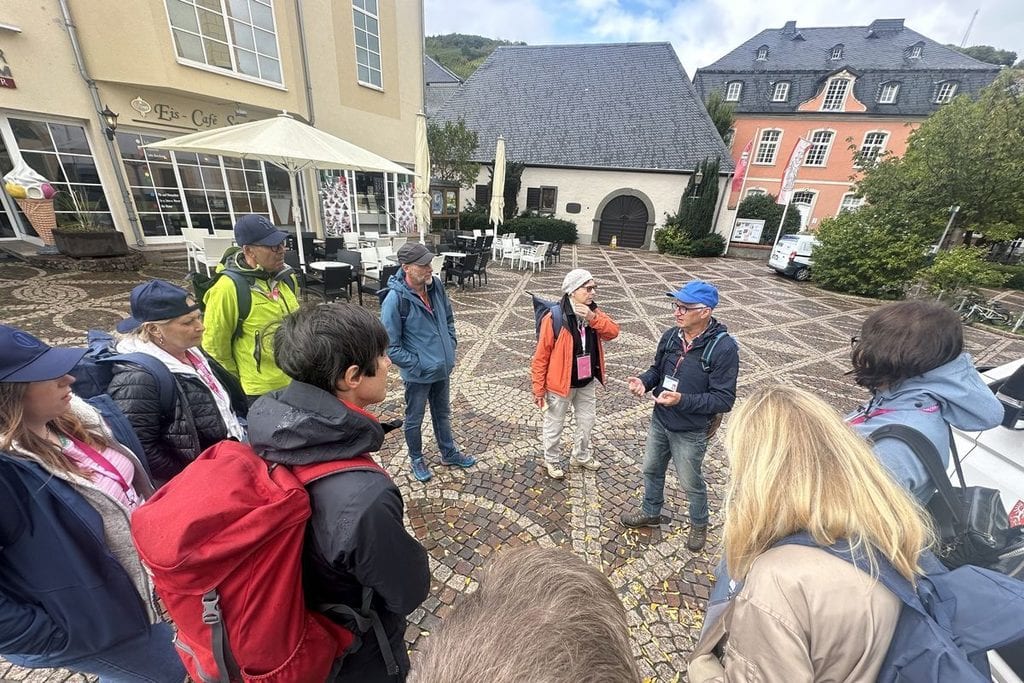 Eine Gruppe von Menschen mit Rucksäcken steht auf einem gepflasterten Platz vor einem Eis-Café und hört einem Mann zu, der in der Mitte spricht.