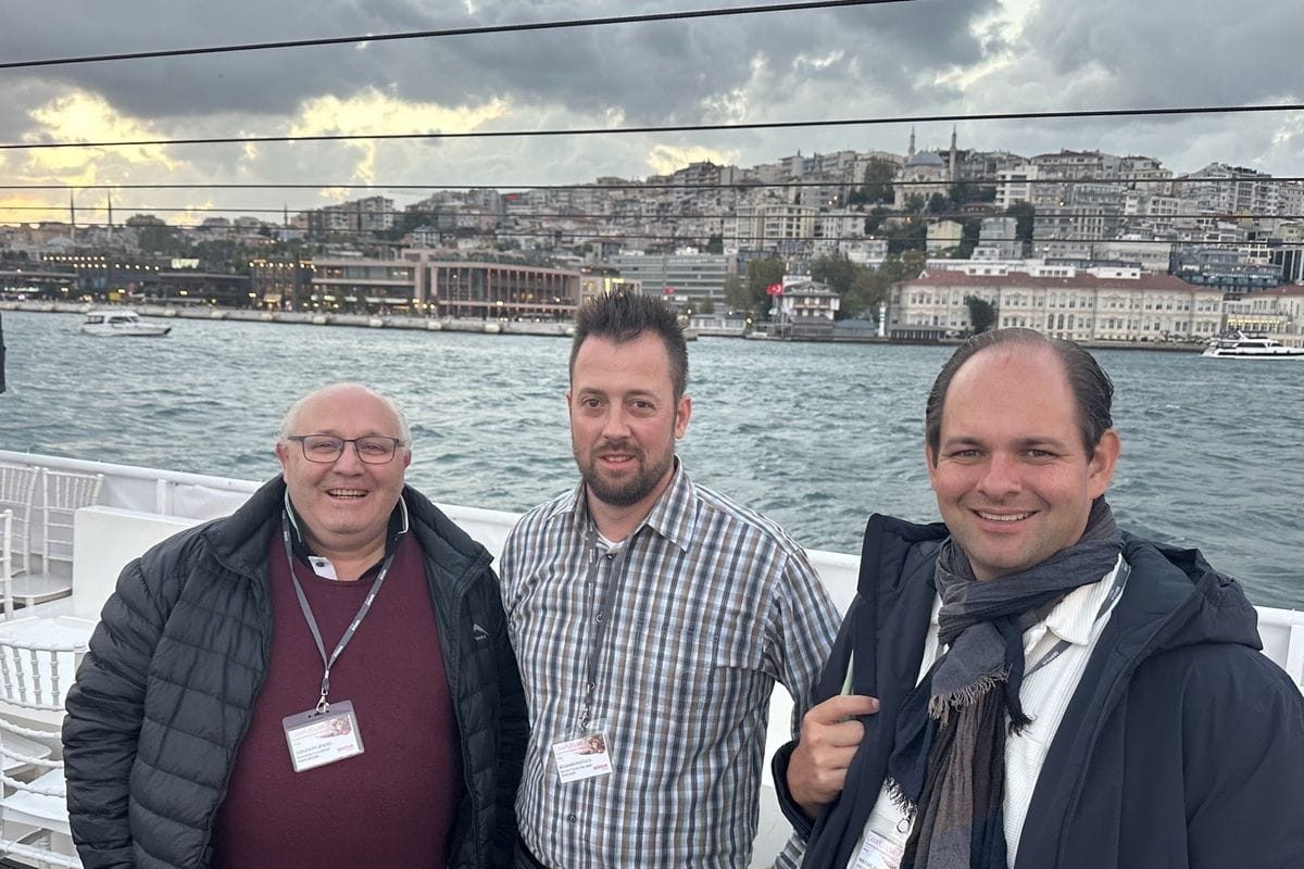 Drei Männer stehen nebeneinander auf einem Schiff vor der Kulisse einer Stadt am Wasser, alle tragen Namensschilder an einem Umhängeband um den Hals.