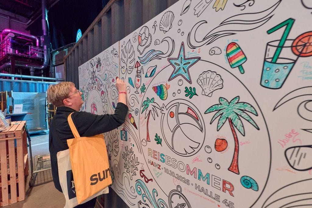 Eine Person mit blonden Haaren und gelber Tragetasche bemalt eine große, mit sommerlichen Motiven bedruckte Wandfläche in einer Industriehalle mit Stiften.