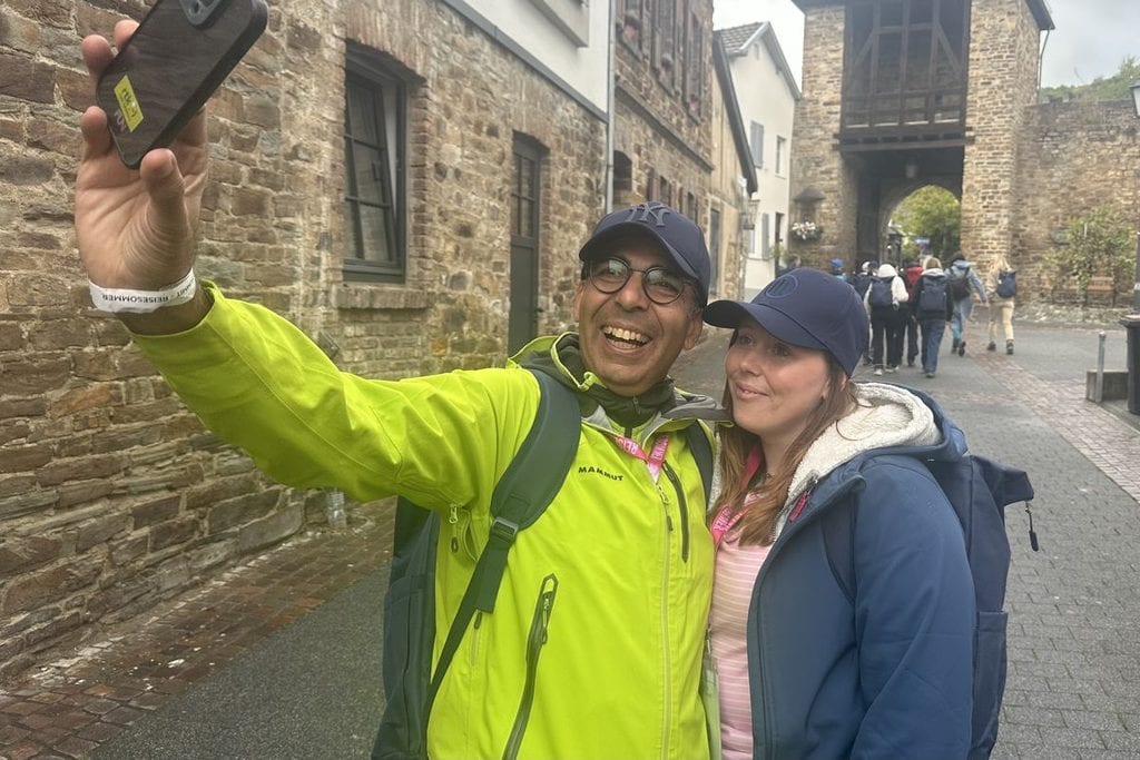 Ein Mann in neongelber Jacke und eine Frau in blauer Jacke machen ein Selfie vor einer historischen Steinmauer mit einem Torbogen in einer Fußgängerzone.