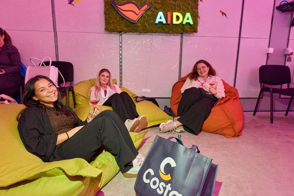 Drei junge Frauen sitzen auf bunten Sitzsäcken vor einer Wand mit AIDA-Logo und halten Weingläser in den Händen, während eine Costa-Tasche im Vordergrund steht.