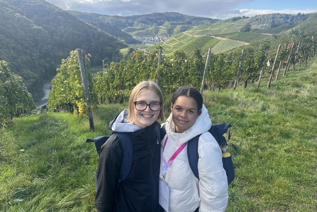 Zwei junge Frauen stehen lächelnd in einem Weinberg vor einer hügeligen Landschaft mit einem Tal und bewaldeten Hängen im Hintergrund bei Tageslicht.