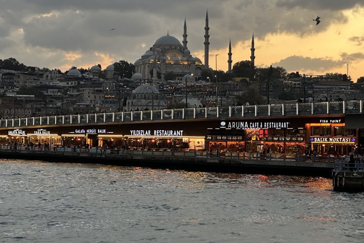Blick auf die beleuchtete Galata-Brücke in Istanbul bei Dämmerung mit der Süleymaniye-Moschee im Hintergrund und dem Wasser des Goldenen Horns im Vordergrund.