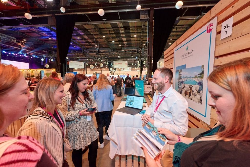 Ein Mann im weißen Hemd steht an einem Messestand von Iberostar und spricht mit drei Frauen, während er Informationsmaterial in den Händen hält.