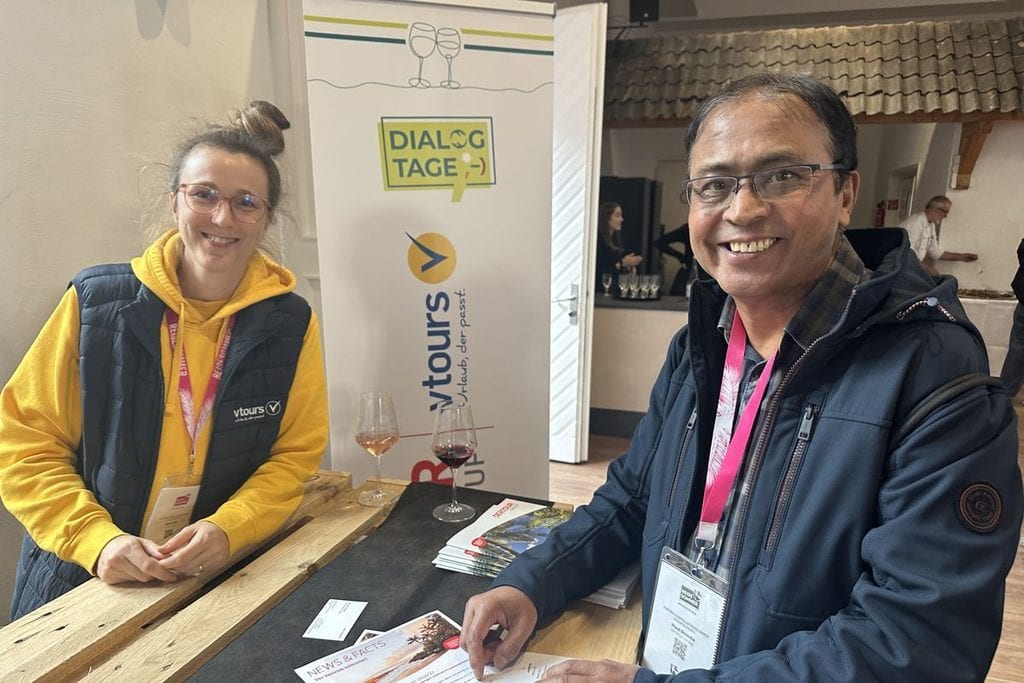 Eine Frau in gelbem Hoodie und ein Mann in blauer Jacke stehen an einem Messestand mit Roll-up-Banner von vtours und Dialogtagen in einem Innenraum.