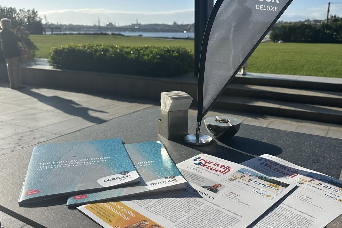 Auf einem Außentisch liegen Broschüren von DERTOUR Deluxe und Ausgaben der Fachzeitschrift touristik aktuell vor einer sonnigen Park- und Wasserlandschaft.