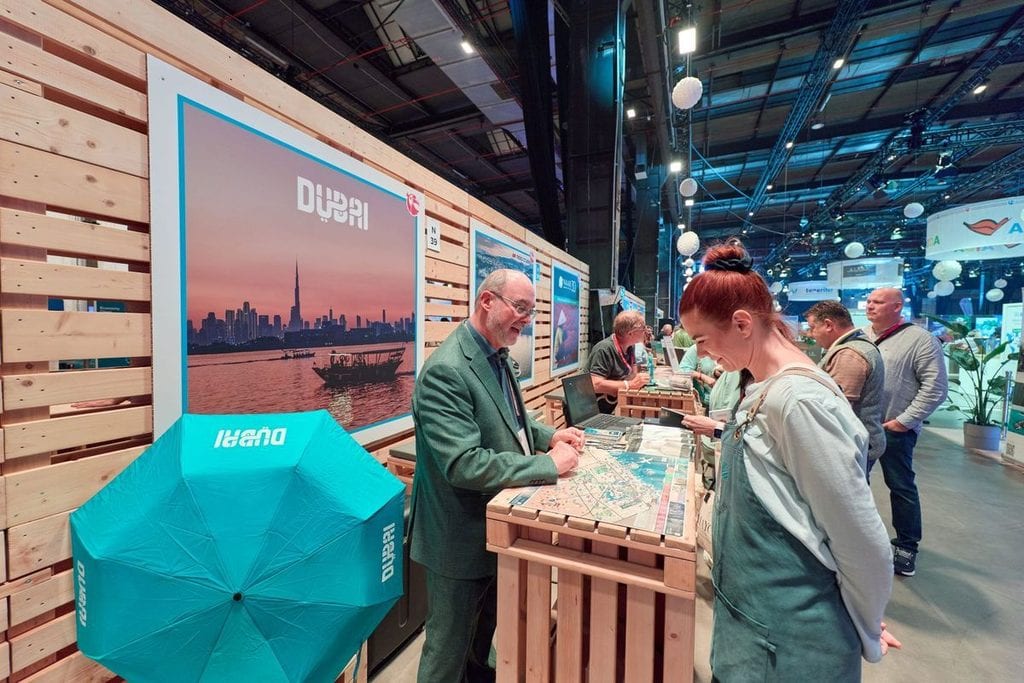 Ein Mann im grünen Anzug und eine Frau unterhalten sich an einem Messestand mit Holzverkleidung, auf dem ein großes Dubai-Plakat und ein Regenschirm sind.