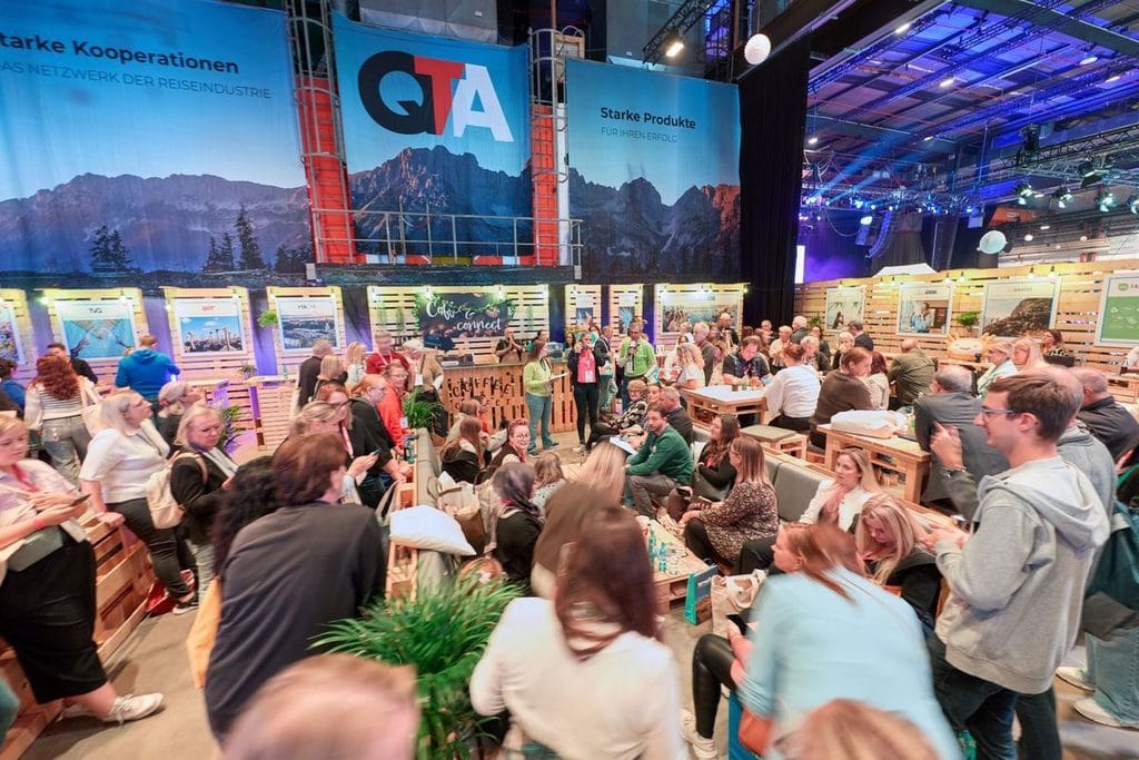 Blick in eine große Messehalle mit vielen Besuchern an Holzständen unter großen Bannern mit QTA-Logo und Bergmotiven vor einer dunklen Deckenkonstruktion.