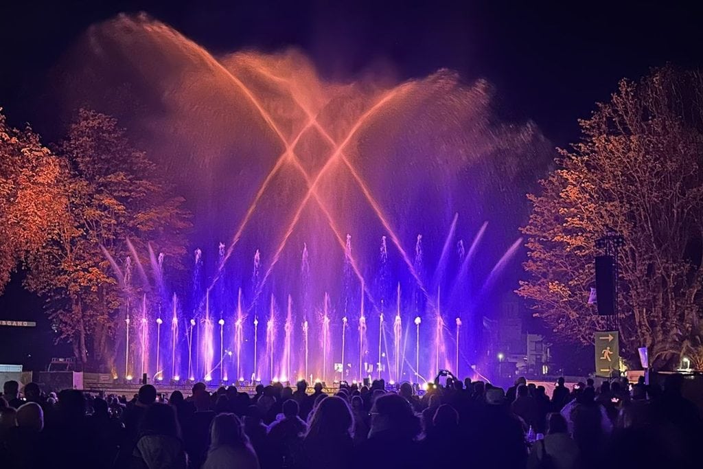 Eine nächtliche Wasserfontänen-Show mit lila und orangefarbener Beleuchtung vor einer Zuschauermenge, die das Spektakel im Freien zwischen Bäumen betrachtet.