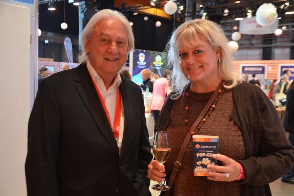 Ein älterer Mann im dunklen Sakko mit rotem Umhängeband und eine blonde Frau in brauner Strickjacke stehen nebeneinander und lächeln in die Kamera.