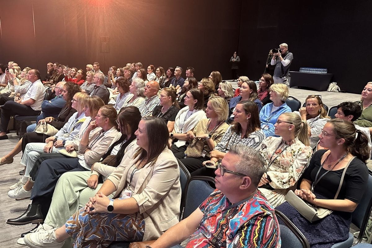 Ein Publikum sitzt in einem abgedunkelten Konferenzsaal auf Stühlen und blickt nach vorne, während ein Fotograf am rechten Bildrand die Szene aufnimmt.