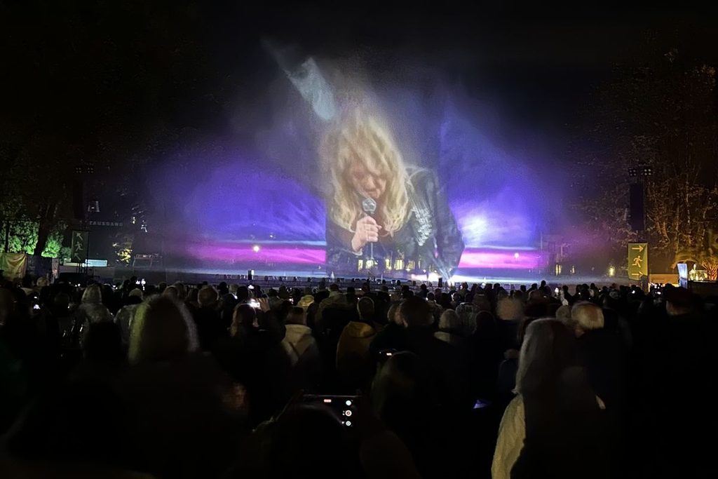 Ein großes Publikum blickt bei Nacht auf eine Wasserleinwand, auf der eine blonde Person mit Mikrofon in violettem Licht projiziert wird. Außenbereich.