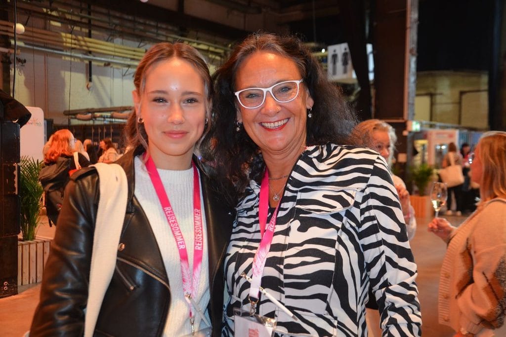 Zwei Frauen stehen nebeneinander in einer Halle und lächeln in die Kamera. Beide tragen pinkfarbene Schlüsselbänder mit der Aufschrift REISESOMMER um den Hals.