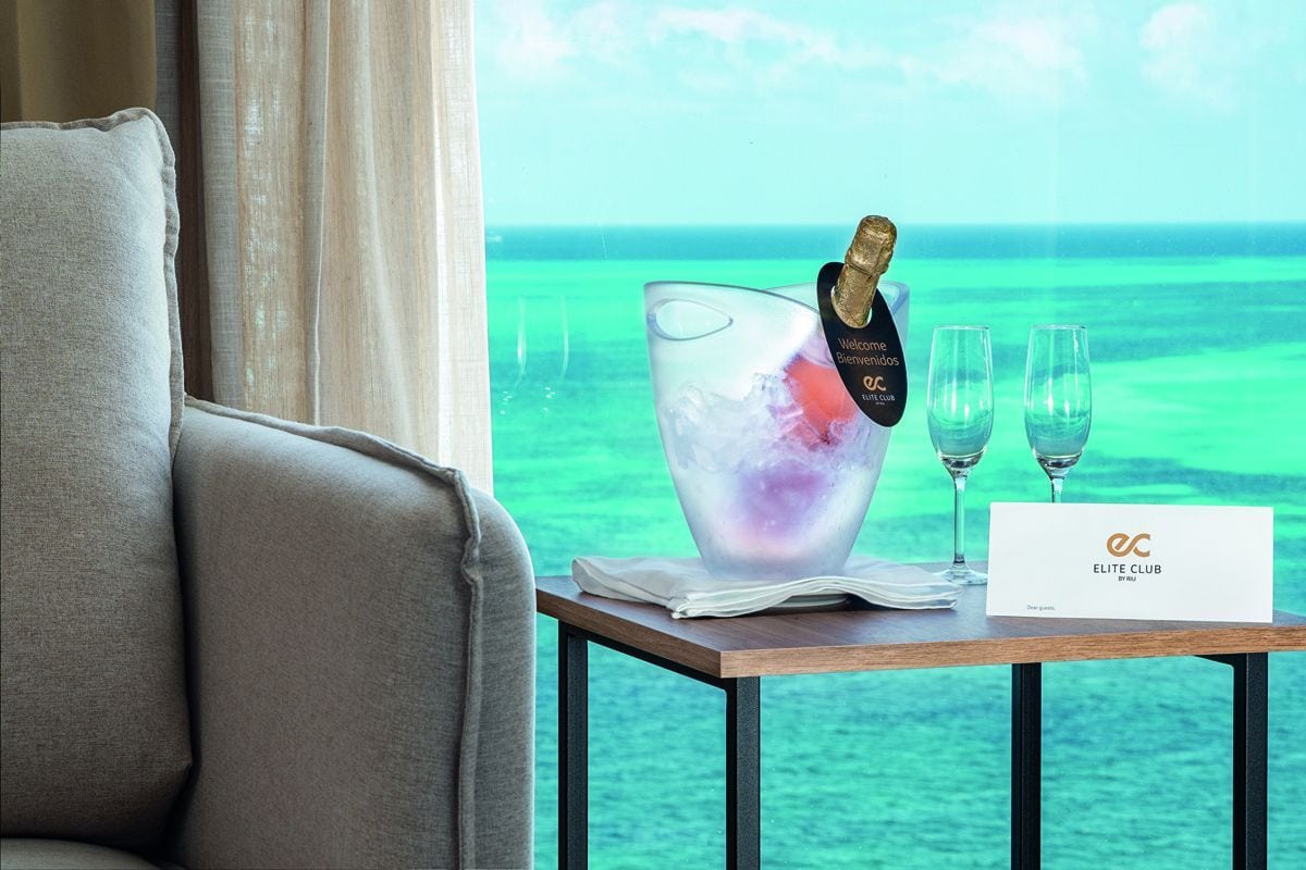 Ein Sektkühler mit Flasche und zwei Sektgläsern steht auf einem Beistelltisch vor einem großen Fenster mit Blick auf das türkisfarbene Meer im Hintergrund.