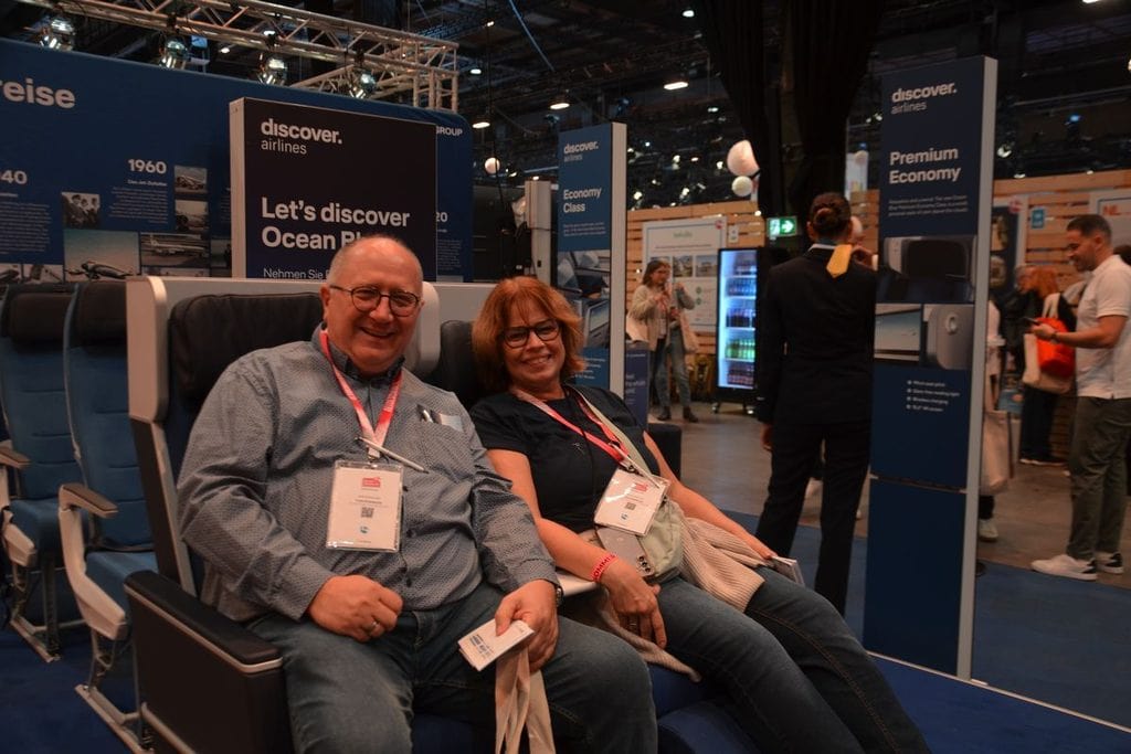 Ein Mann und eine Frau sitzen lächelnd in Flugzeugsitzen auf einer Messe, im Hintergrund sind Infoständer von discover airlines und Messebesucher zu sehen.