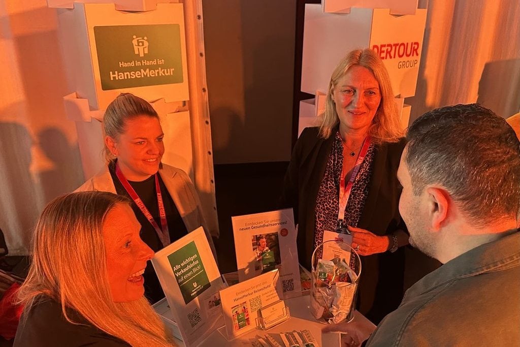 Drei Frauen und ein Mann stehen an einem Messestand von HanseMerkur und DERTOUR Group und führen ein Gespräch vor aufgestellten Informationsaufstellern.