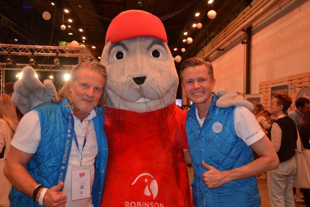 Zwei Männer in blauen Westen stehen neben einem großen Robby-Maskottchen mit roter Kappe und rotem T-Shirt in einer hell erleuchteten Messehalle zusammen.