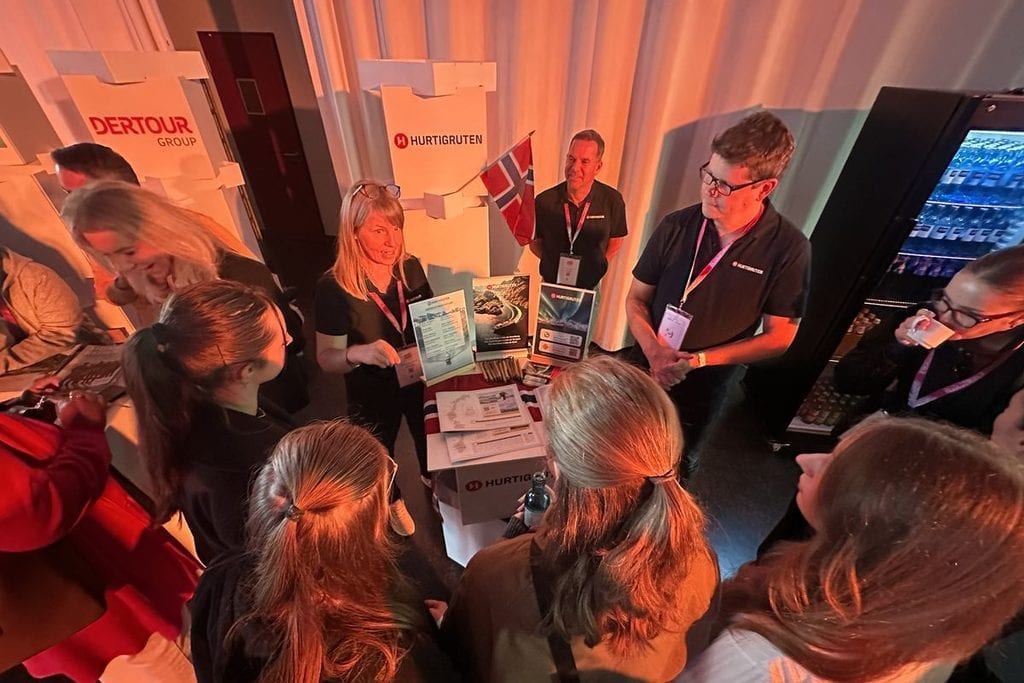 Eine Gruppe von Menschen versammelt sich um einen Messestand von Hurtigruten, an dem Informationsmaterialien und eine norwegische Flagge zu sehen sind.