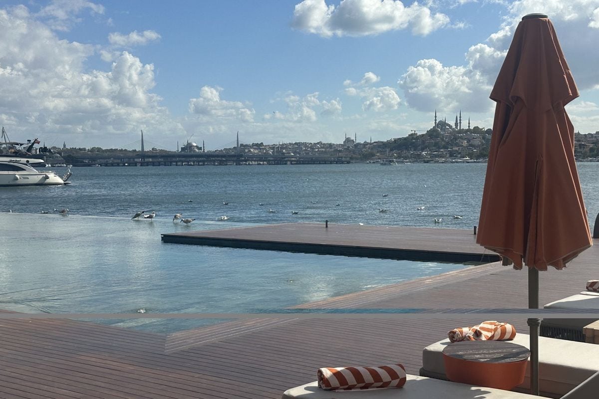 Blick von einer Holzterrasse mit Sonnenschirm und Liegen auf das Wasser, mit Vögeln im Vordergrund und der Skyline von Istanbul im sonnigen Hintergrund.