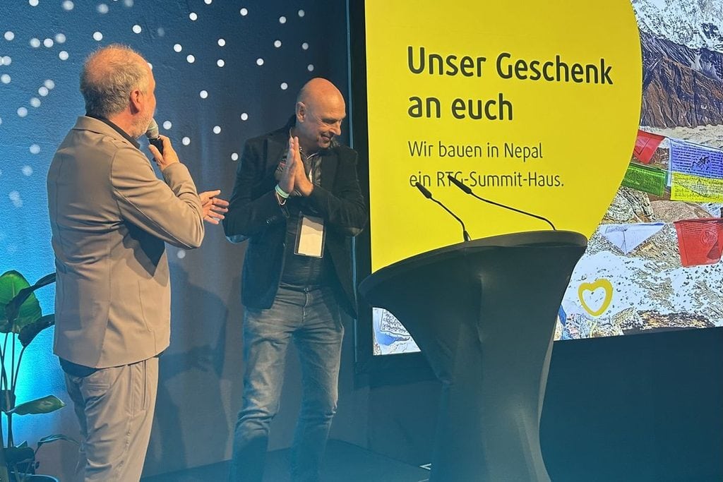 Zwei Männer stehen auf einer Bühne vor einer gelben Leinwand mit Text und Bergmotiv. Einer spricht in ein Mikrofon, der andere klatscht in die Hände.