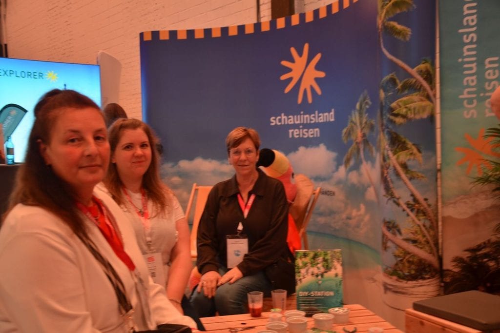 Drei Frauen stehen vor einem Messestand von schauinsland-reisen mit Palmenmotiv, einer DIY-Station auf einem Holztisch und einem ausgestellten Flamingo.