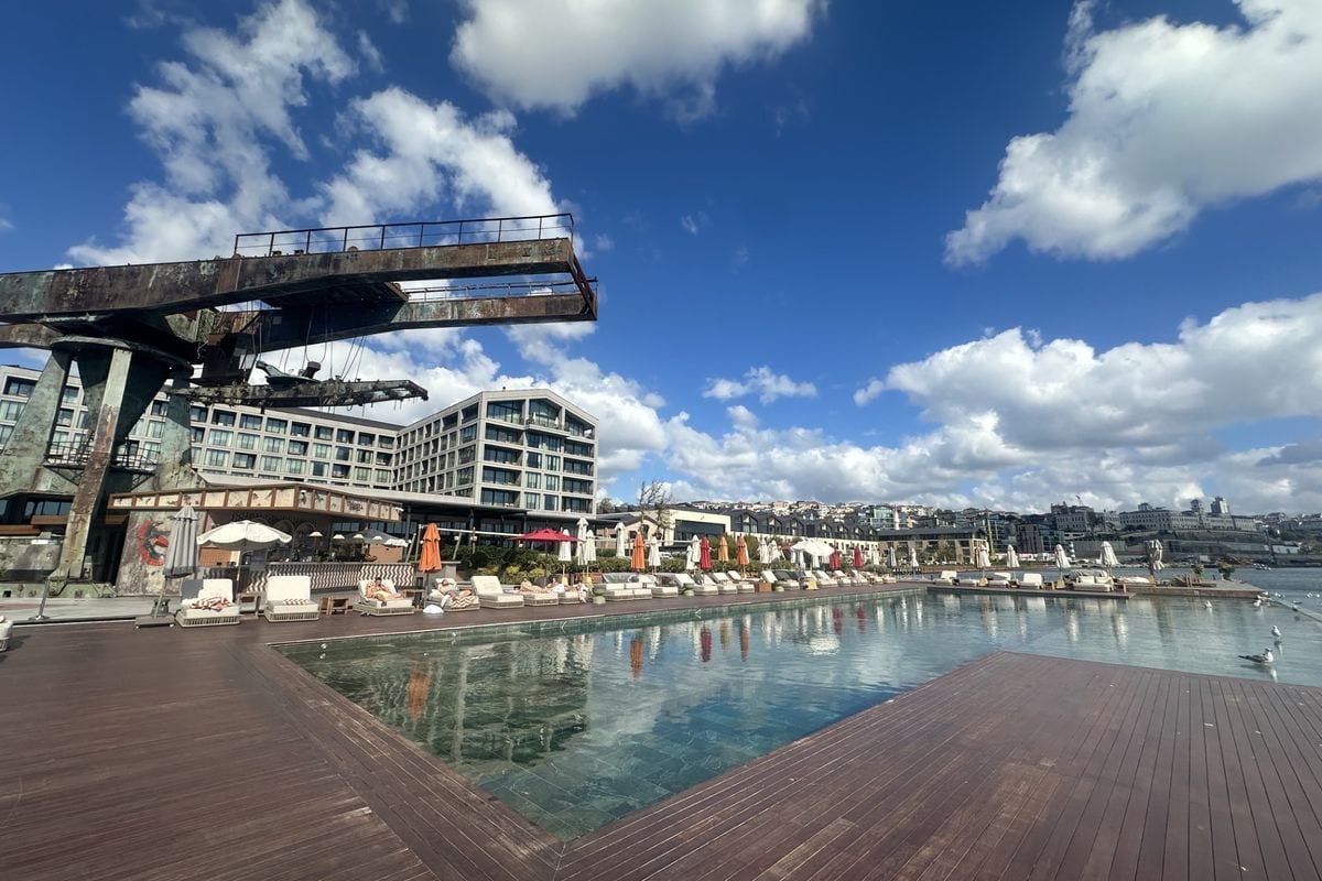 Ein Außenpool mit Holzdeck und Liegestühlen vor einem Hotelgebäude, über dem ein großer, rostiger Industriekran unter einem bewölkten Himmel aufragt.
