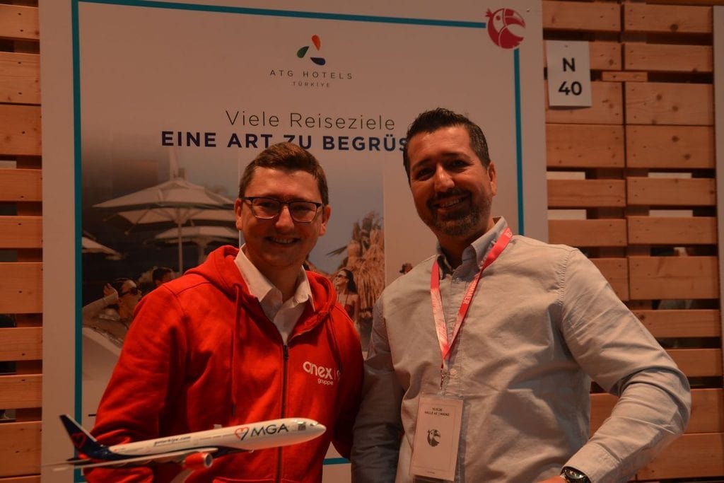 Zwei lächelnde Männer stehen vor einem Werbeplakat von ATG Hotels. Der linke Mann trägt einen roten Kapuzenpullover und hält ein kleines Flugzeugmodell.