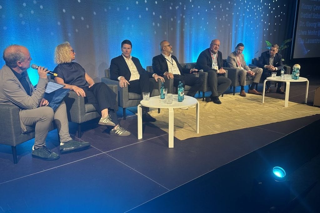 Sechs Personen sitzen in einer Reihe auf Sesseln auf einer Bühne vor einer blauen Wand mit Lichtpunkten und diskutieren an zwei runden weißen Tischen.