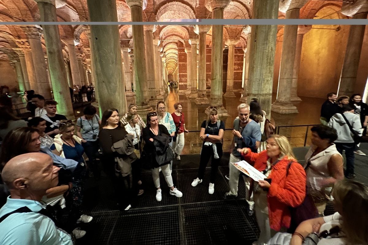 Eine Gruppe von Menschen steht auf einem Metallsteg in der Basilica Cisterna in Istanbul zwischen zahlreichen antiken Säulen und hört einer Frau zu.