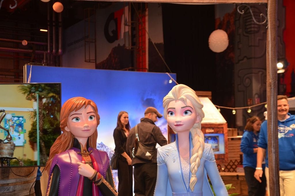 Zwei lebensgroße Figuren von Anna und Elsa aus dem Film Die Eiskönigin stehen in einer Messehalle vor einem blauen Hintergrund mit weiteren Personen.