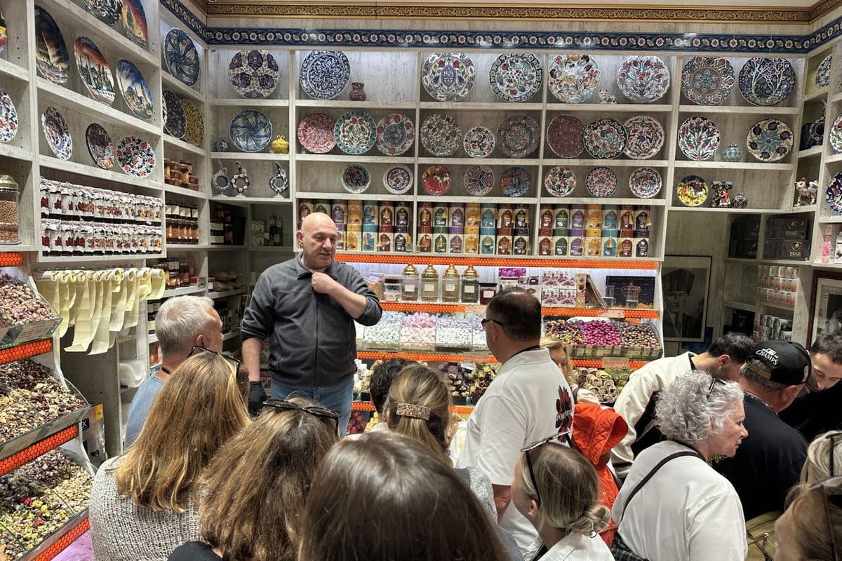 Ein Mann spricht vor einer Gruppe von Menschen in einem Geschäft, das mit kunstvoll bemalten Tellern und verschiedenen Waren in Regalen gefüllt ist.