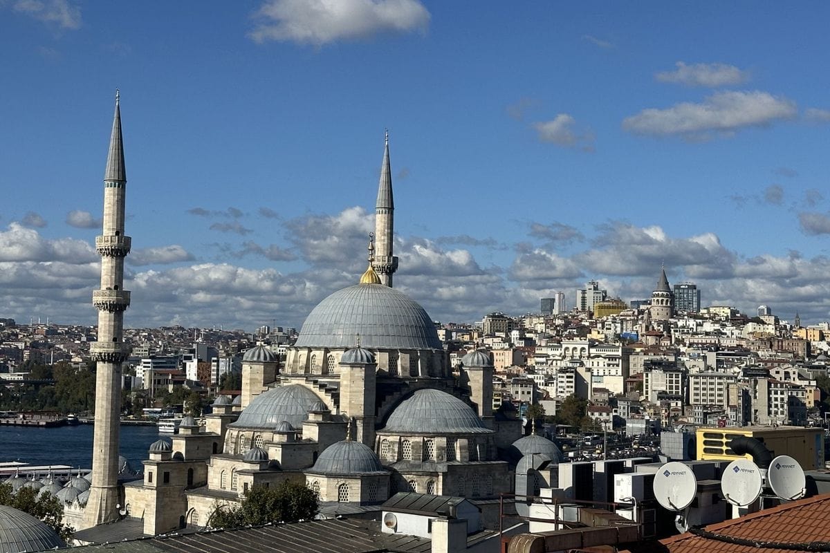 Blick auf die Yeni-Moschee in Istanbul mit ihren Kuppeln und Minaretten vor einer dichten Stadtkulisse mit dem Galata-Turm unter einem blauen Himmel.