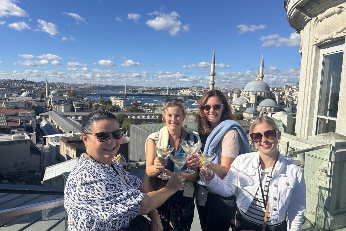 Vier Frauen stehen auf einer Dachterrasse, halten Martinigläser in die Höhe und lächeln vor einer Skyline mit Moscheen und dem Goldenen Horn in Istanbul.