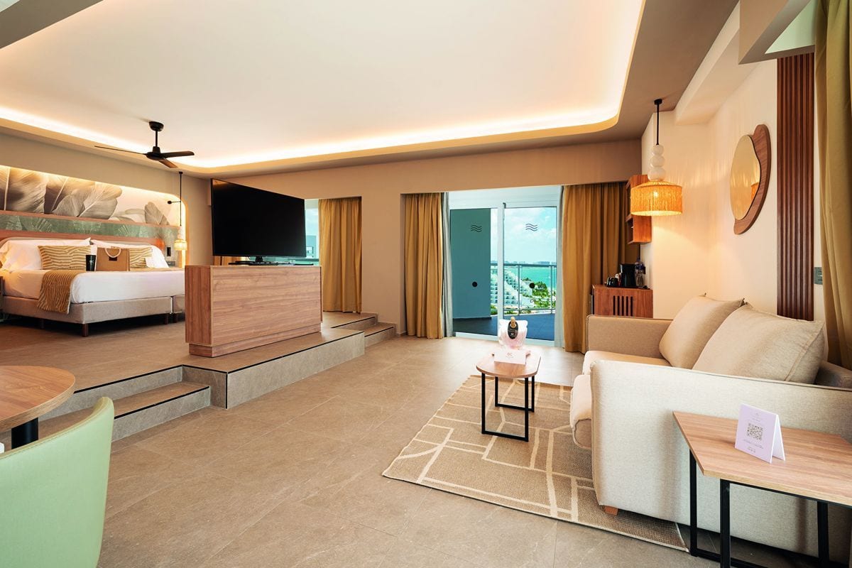 Ein modernes Hotelzimmer mit erhöhtem Schlafbereich, großem Flachbildfernseher, gemütlichem Sofa, Couchtisch und Blick auf einen Balkon mit Meerblick.