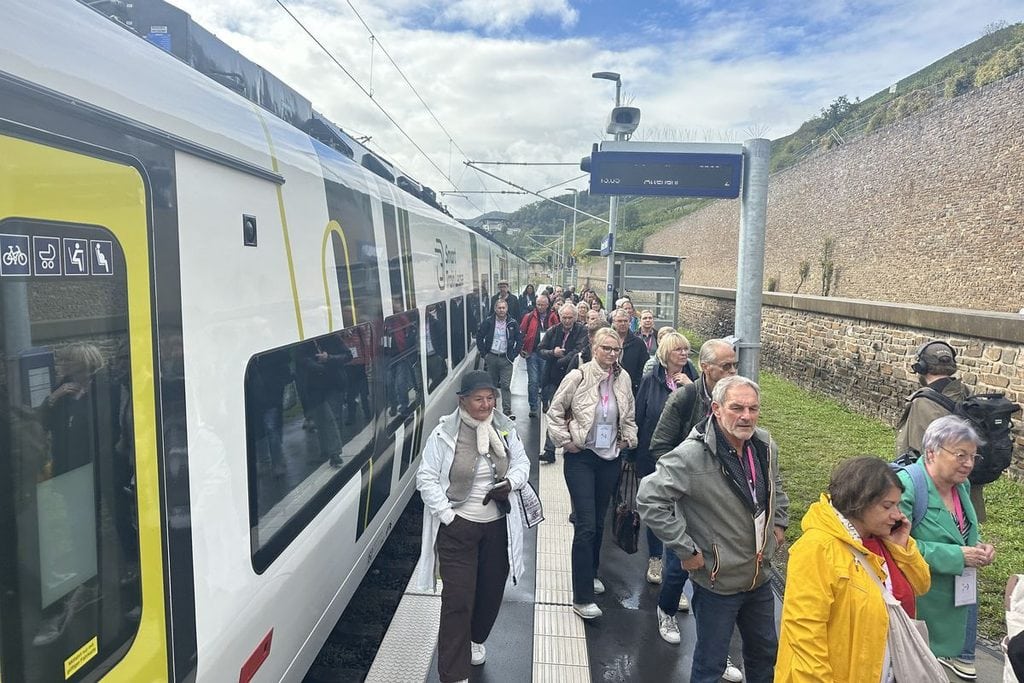 Eine Gruppe von Menschen steigt an einem Bahnsteig aus einem modernen Zug aus, der neben einer hohen Steinmauer und einem digitalen Anzeigeschild hält.