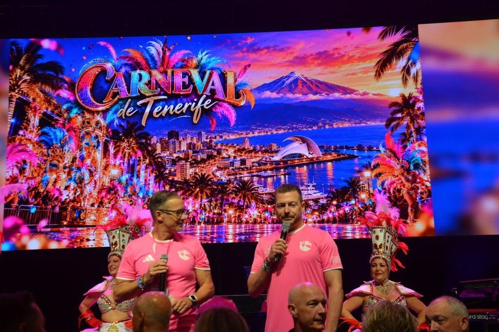 Zwei Männer in rosa T-Shirts stehen mit Mikrofonen auf einer Bühne vor einer großen Leinwand, die den Carneval de Tenerife und eine Küstenstadt zeigt.