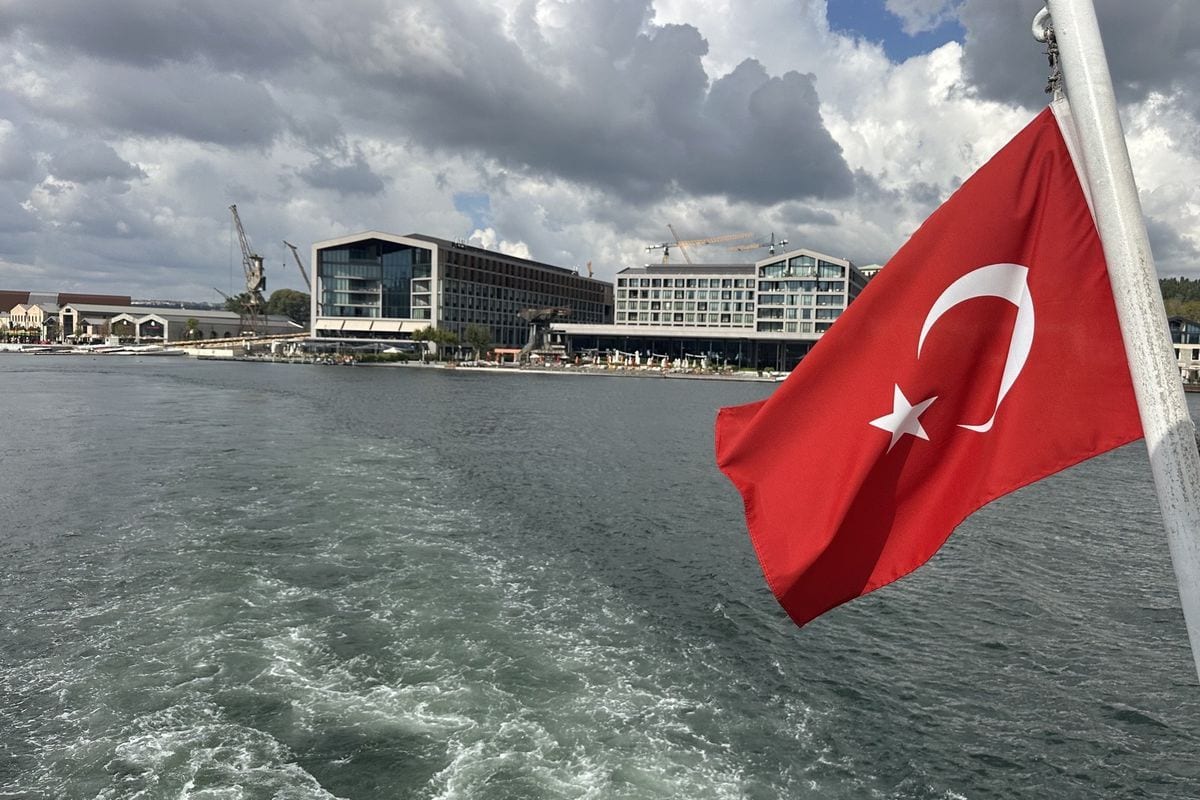 Eine türkische Flagge weht an einem Mast auf einem Schiff vor einer Küstenlinie mit modernen Hotelgebäuden und Kränen unter einem bewölkten Himmel.