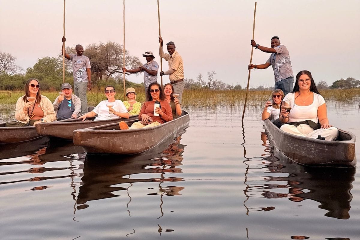 Eine Gruppe von Menschen sitzt in zwei traditionellen Einbäumen auf einem ruhigen Gewässer, während Männer die Boote mit langen Stangen durch das Wasser führen.