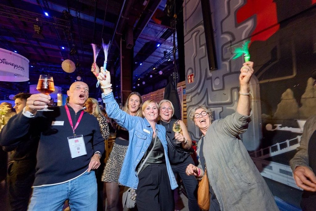 Eine Gruppe von Menschen in einer festlich beleuchteten Halle hebt in fröhlicher Stimmung Gläser und leuchtende Accessoires in die Höhe, um zu feiern.