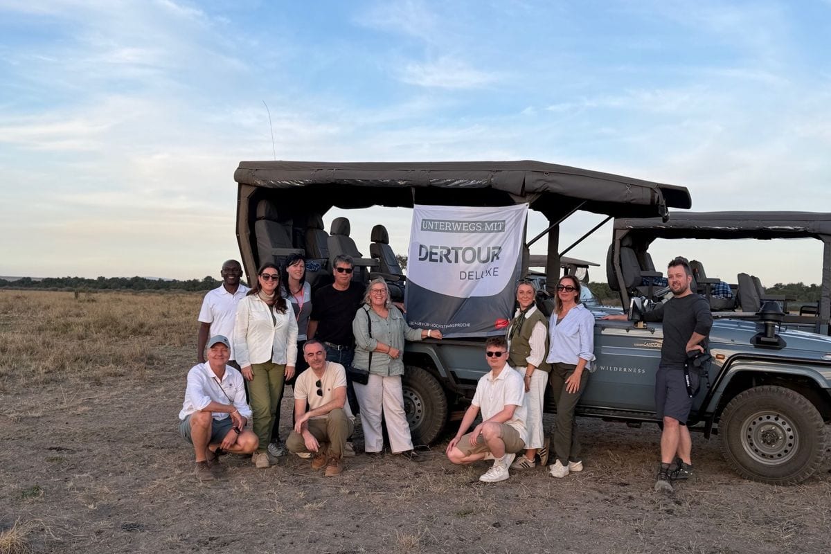 Eine Gruppe von elf Personen posiert vor einem offenen Geländewagen in einer Savannenlandschaft, an dem ein Banner mit der Aufschrift Dertour Deluxe hängt.